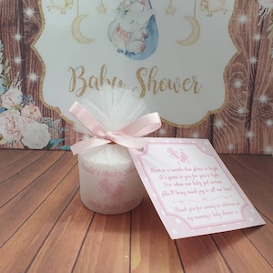 Puede incluir: Una peque&ntilde;a vela blanca con una cinta rosa y un lazo de tul. La vela est&aacute; decorada con huellas de pies rosas y el texto "Paula's Baby Shower 20th June 2020". Una tarjeta rosa con lunares blancos y el texto "Aqu&iacute; tienes una vela que brilla tanto Te la damos para que la enciendas For when our baby girl arrives She'll bring much joy to all our lives! Gracias por venir a celebrar en mi baby shower xx" est&aacute; unida a la vela.