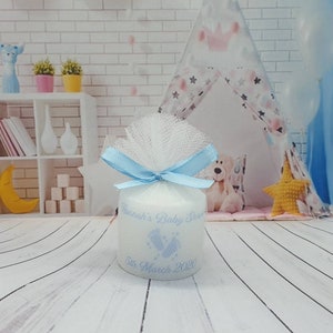 Puede incluir: Una vela blanca con una cinta azul y tul. La vela est&aacute; decorada con un dise&ntilde;o de huella azul y el texto "Hannah's Baby Shower 5th March 2021".