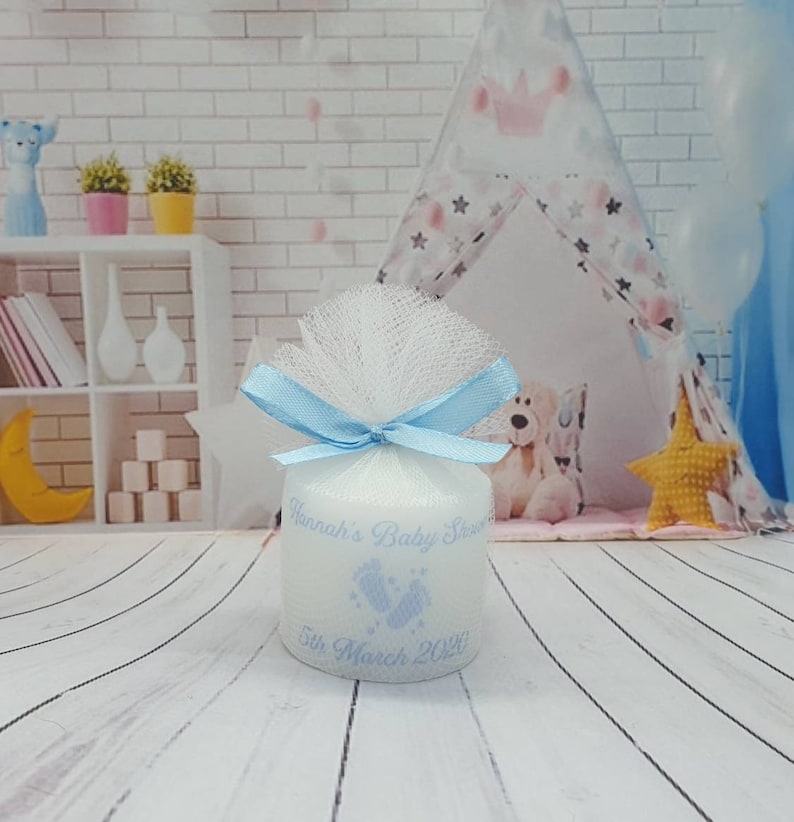 Puede incluir: Una vela blanca con una cinta azul y tul. La vela est&aacute; decorada con un dise&ntilde;o de huella azul y el texto "Hannah's Baby Shower 5th March 2021".
