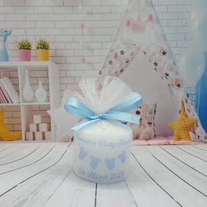 Puede incluir: Una vela blanca con una cinta azul y un lazo de tul. La vela est&aacute; decorada con un dise&ntilde;o azul y blanco y el texto "[Name]'s Baby Shower 5th March 2020".