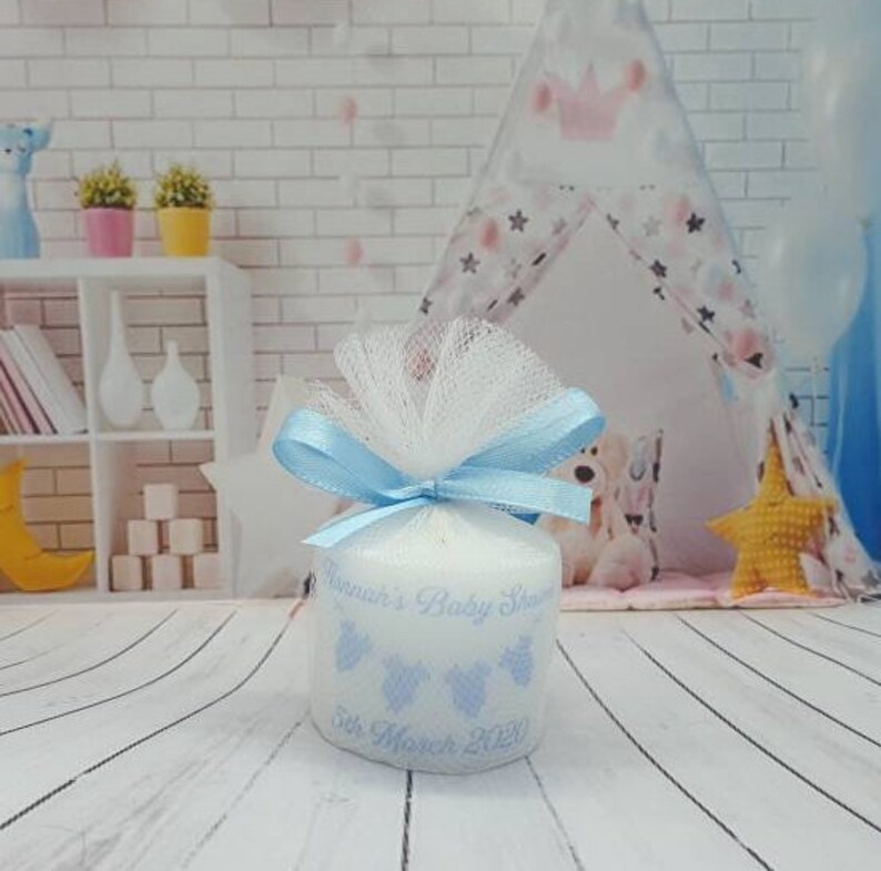 Puede incluir: Una vela blanca con una cinta azul y un lazo de tul. La vela est&aacute; decorada con un dise&ntilde;o azul y blanco y el texto "[Name]'s Baby Shower 5th March 2020".