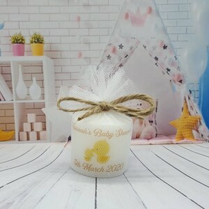 Puede incluir: Una vela blanca con un dise&ntilde;o amarillo de un beb&eacute; y un pato. La vela est&aacute; envuelta en tul blanco y atada con un lazo de cuerda marr&oacute;n. La vela tiene el texto "Hannah's Baby Shower 5th March 2020" impreso en ella.