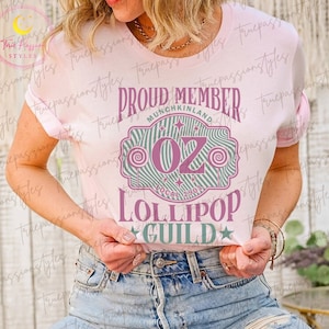 May include: Light pink t-shirt with a graphic featuring the text "PROUD MEMBER MUNCHKINLAND OZ LOLLIPOP GUILD" in purple and green. The design includes stars and lollipop imagery.