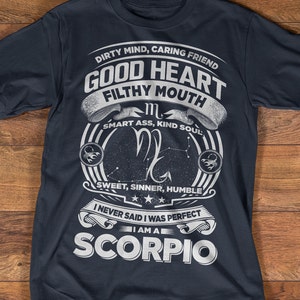 May include: A dark blue t-shirt with a white graphic design featuring the zodiac sign Scorpio. The graphic includes the text "Dirty Mind, Caring Friend, Good Heart, Filthy Mouth, Smart Ass, Kind Soul, Sweet, Sinner, Humble, I Never Said I Was Perfect, I Am A Scorpio".