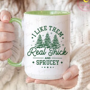Könnte beinhalten: Weiße Keramik-Tasse mit grünem Innenraum und Griff. Die Tasse zeigt den Text "I LIKE THEM Real Thick AND Sprucey" in Grün, zusammen mit Illustrationen von drei Weihnachtsbäumen und Schneeflocken. Die Tasse wird von einer Person gehalten, die einen weißen Pullover trägt.