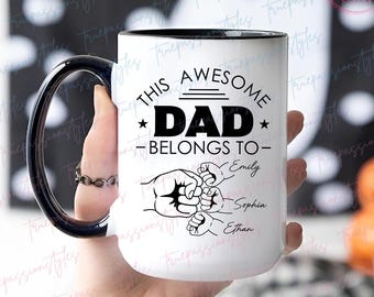 Może przedstawiać: Biały ceramiczny kubek z czarnym uchwytem i obramowaniem. Kubek zawiera napis "THIS AWESOME DAD BELONGS TO" z imionami Emily, Sophia i Ethan pod grafiką zaciśniętych pięści. Kubek jest trzymany przez osobę.