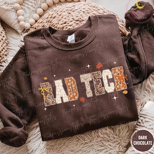 May include: Dark chocolate-colored sweatshirt with the text "BAD TECH" in a patchwork design. The letters are decorated with floral and autumn-themed patterns, with small star and leaf accents. The sweatshirt has a crew neck and long sleeves.