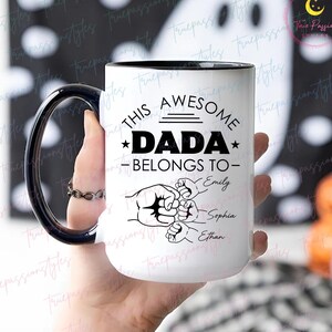 Könnte beinhalten: Weißer Keramikbecher mit schwarzem Henkel und Rand. Der Becher trägt den Text "THIS AWESOME DADA BELONGS TO" mit den Namen Emily, Sophia und Ethan. Eine Faust-Grafik ist ebenfalls enthalten.