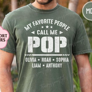 May include: Moss green t-shirt with the text "MY FAVORITE PEOPLE CALL ME POP" in white. Below the word "POP" are the names Olivia, Noah, Sophia, Liam, and Anthony. The shirt has a crew neck and short sleeves.