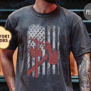 Puede incluir: Una camiseta gris oscuro con un diseño de bandera estadounidense desgastado que presenta a un lineman trabajando en un poste de energía. El texto "PEPPER" es visible en la esquina superior derecha de la imagen.