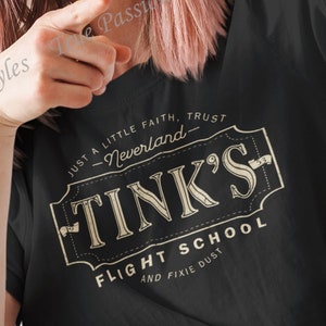 May include: A black t-shirt featuring a vintage-style logo that reads "Just a little faith, trust - Neverland - Tink's Flight School and Pixie Dust".