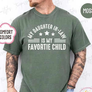 May include: A moss green t-shirt with the white text "MY DAUGHTER IN-LAW IS MY FAVORITE CHILD". The shirt has a crew neck and short sleeves. The words "COMFORT COLORS" are in a pink bubble on the left side of the shirt.
