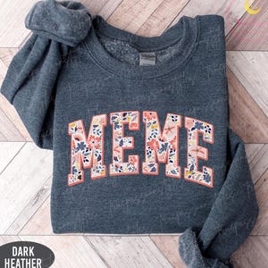 May include: Dark heather gray sweatshirt with the word "MEME" in a floral design. The letters are outlined in a coral color, and the floral pattern includes pink, blue, and white flowers. The sweatshirt is folded on a wooden surface.