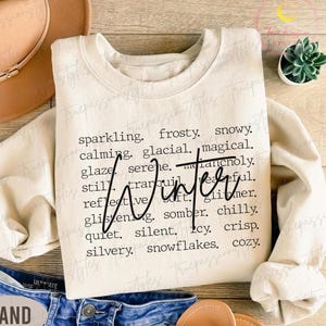 May include: Cream-colored sweatshirt with the word "Winter" in a cursive font. Surrounding the word are descriptive words like "sparkling," "frosty," and "snowy." The sweatshirt is paired with blue denim jeans and a tan hat.