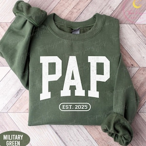 May include: Military green sweatshirt with the word "PAP" in large, distressed white letters. Below the word "PAP" is the text "EST. 2025" in a white, rounded rectangle. The sweatshirt has a crew neck and long sleeves.