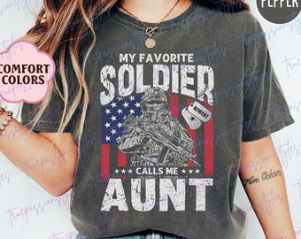 Può includere: T-shirt grigio scuro con la scritta "MY FAVORITE SOLDIER CALLS ME AUNT" e un'illustrazione di un soldato con un fucile. La bandiera americana è sullo sfondo. Il nome "ROBERT" è su una targhetta.