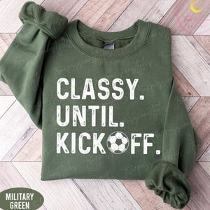 May include: Military green sweatshirt with the text "CLASSY. UNTIL. KICKOFF." in white. A soccer ball graphic is below the text. The sweatshirt is displayed on a light brown wooden surface.