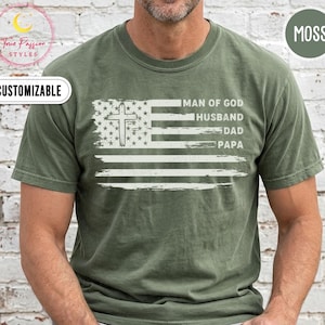May include: A moss green short-sleeved t-shirt featuring a white graphic of an American flag with a cross integrated into the star field. The text MAN OF GOD, HUSBAND, DAD, and PAPA is printed in white on the right side of the flag graphic.