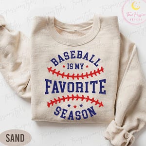 May include: A sand-coloured sweatshirt featuring the text "BASEBALL IS MY FAVORITE SEASON" in blue and red. The design includes baseball stitching and stars. The sweatshirt has long sleeves and a crew neck.