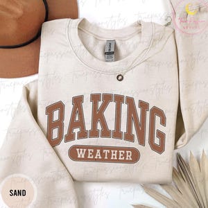 Op de afbeelding: Zandkleurige sweatshirt met de tekst "BAKING WEATHER" in bruine blokletters. De sweatshirt heeft een ronde hals en lange mouwen. Een bruine hoed en een ketting met een hanger zijn zichtbaar. De sweatshirt is van het merk Gildan.