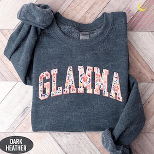 May include: Dark heather gray sweatshirt with the word "GLAMMA" in a floral design. The letters are outlined in a peach color, and the floral pattern includes pink, blue, and white flowers. The sweatshirt is folded on a wooden surface.