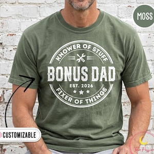 May include: A moss green short-sleeve t-shirt featuring a white circular graphic with the text "KNOWER OF STUFF", "BONUS DAD", "EST. 2026", and "FIXER OF THINGS" surrounding a crossed wrench and screwdriver icon and three stars.