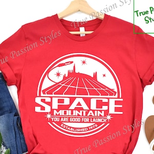 Może przedstawiać: Czerwona koszulka z białym nadrukiem logo Space Mountain. Logo przedstawia biały statek kosmiczny, górę i tekst "Space Mountain", "You are good for launch" oraz "Established 1975".
