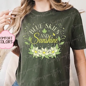 May include: A green t-shirt with the text "Blue Skies and Sunshine" in a white and yellow font. The text is surrounded by a floral design with white flowers and green leaves.