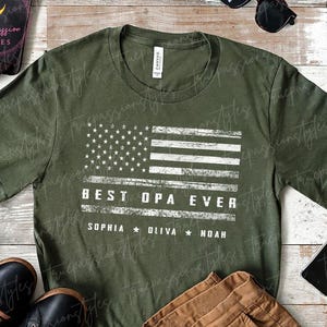 May include: Heather military green t-shirt with a distressed American flag design. The text reads "BEST OPA EVER" with the names Sophia, Oliva, and Noah below. The shirt is displayed with brown pants and black shoes.