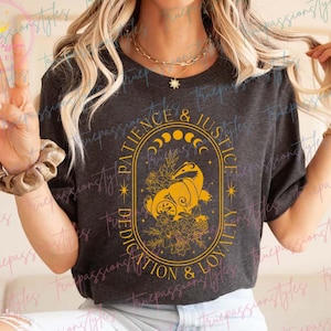 May include: A dark gray t-shirt with a yellow graphic design featuring a crescent moon, stars, and a floral design. The text on the graphic reads "Patience & Justice, Dedication & Loyalty".