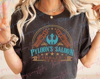 May include: A dark grey t-shirt with a graphic design featuring a stylised image of a spaceship with the text "Pylon's Saloon" and "Rambler's Reach" underneath.