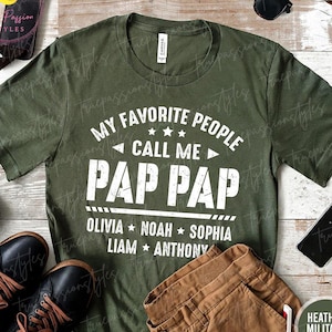May include: Heather military green t-shirt with the text "MY FAVORITE PEOPLE CALL ME PAP PAP" in white. The names Olivia, Noah, Sophia, Liam, and Anthony are listed below. The shirt is displayed with brown pants and black shoes.