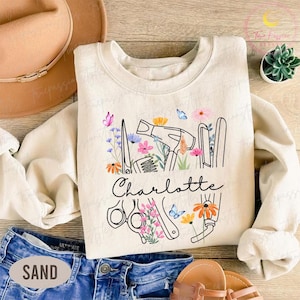 May include: A cream-coloured sweatshirt with a floral design incorporating scissors, a hairdryer, and other salon tools. The name "Charlotte" is written in cursive script below the design. The sweatshirt is paired with blue denim shorts.