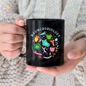 Puede incluir: Taza de caf&eacute; negra con un dise&ntilde;o circular blanco con el texto "MEOWCROBIOLOGY" y gatos y bacterias de dibujos animados de colores.