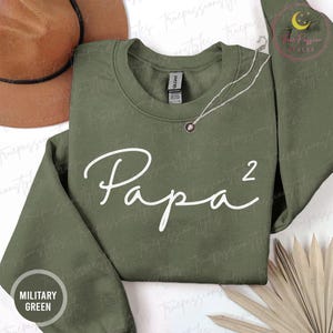 Op de afbeelding: Een olijfgroene sweatshirt met het woord "Papa²" in witte letters. Een zilveren ketting met een kleine hanger is over het shirt gedrapeerd. De woorden "Military Green" staan in een cirkel linksonder.