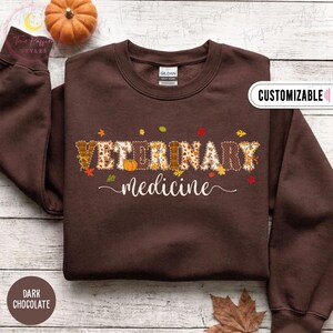 Puede incluir: Una sudadera de cuello redondo en color marrón chocolate oscuro con el texto VETERINARY en letras grandes con un patrón otoñal de calabazas y hojas, seguido de la palabra medicine en una fuente cursiva blanca en el pecho.