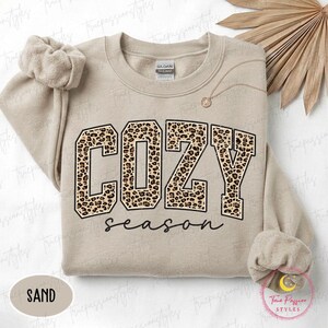 May include: A sand-colored crewneck sweatshirt featuring the word COZY in large block letters with a leopard print pattern and the word season written in black cursive script below. The garment has a Gildan brand label at the inner neckline.