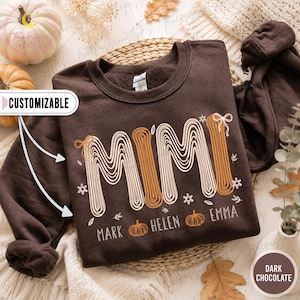 Könnte beinhalten: Ein dunkel schokoladenbraunes Sweatshirt mit Rundhalsausschnitt, das das Wort MIMI in einer stilisierten, mehrzeiligen Regenbogen-Schriftart mit kleinen Schleifen zeigt. Unter dem Text stehen die Namen MARK, HELEN und EMMA, getrennt durch Kürbisse.