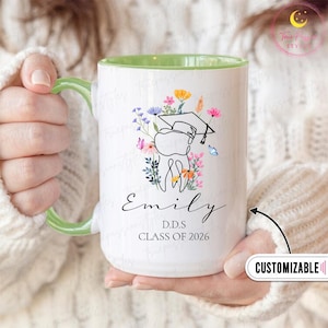 May include: White ceramic mug with a green handle and interior. The mug features a floral design with a graduation cap and a tooth illustration. The name "Emily", "D.D.S", and "Class of 2026" are printed on the mug. The mug is customizable.
