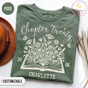May include: A moss green t-shirt featuring a white graphic of an open book with a bouquet of wildflowers emerging from the pages. The text reads Chapter Twenty, 20, 06, and Charlotte. A thin gold chain necklace rests on the fabric near the collar.