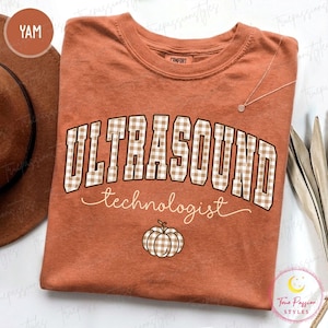 May include: A rust-colored short-sleeve t-shirt featuring the text ULTRASOUND in a beige and white gingham pattern, with the word technologist in white cursive script below it, and a small gingham pumpkin graphic at the bottom center.