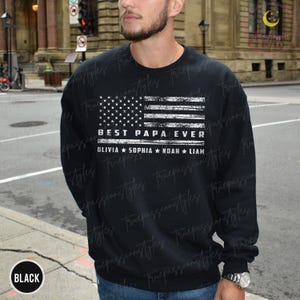 K&ouml;nnte beinhalten: Schwarzes Sweatshirt mit einem verblassten amerikanischen Flaggen-Design. Der Text "BEST PAPA EVER" wird unter der Flagge angezeigt, mit den Namen OLIVIA, SOPHIA, NOAH und LIAM darunter. Das Wort "BLACK" befindet sich in einem Kreis unten.