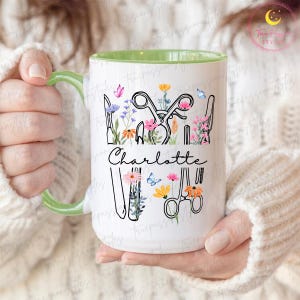 Op de afbeelding: Witte keramische mok met een groene handgreep en binnenkant. De mok heeft een bloemenontwerp met scharen, kammen en de naam "Charlotte" in script. Vlinders en bloemen in roze, geel en blauw versieren de mok.