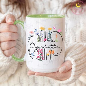 May include: White ceramic mug with a green handle and interior, featuring the name "Charlotte" in a black script font. The mug is decorated with colourful flowers and butterflies, creating a floral design.