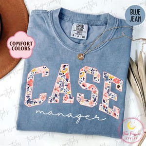 Könnte beinhalten: Blaues Comfort Colors T-Shirt mit der Aufschrift "CASE manager" in einem floralen Design. Eine goldene Halskette mit Anhänger ist über dem Shirt drapiert. Das Shirt hat einen Rundhalsausschnitt und kurze Ärmel.