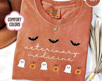 May include: A burnt orange t-shirt with the words "veterinary medicine" in white script. The shirt features black bat graphics above the text and ghost and pumpkin graphics below. A gold necklace is draped on the shirt.