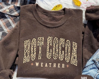 May include: A dark chocolate brown sweatshirt featuring the text "HOT COCOA WEATHER" in a leopard print design. The crew neck sweatshirt has long sleeves. The words "DARK CHOCOLATE" are in a brown oval at the bottom right.