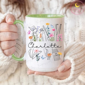 Może przedstawiać: Biały ceramiczny kubek z zielonym uchwytem i wnętrzem, z kwiatowym wzorem z narzędziami do rzęs i imieniem "Charlotte" w czarnym skrypcie. Projekt zawiera motyle i kwiaty.