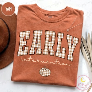 May include: A rust-colored short-sleeve t-shirt featuring the text "Early intervention" in a large, blocky, beige and white gingham pattern. Below the text is a small, matching gingham pumpkin graphic. The shirt has a crew neckline.