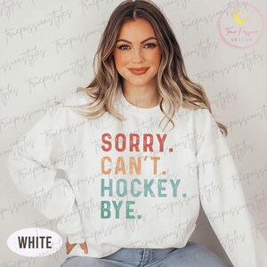 May include: White sweatshirt with the text "SORRY. CAN'T. HOCKEY. BYE." in a retro font. The sweatshirt has a crew neck and long sleeves. The word "WHITE" is at the bottom left of the sweatshirt.